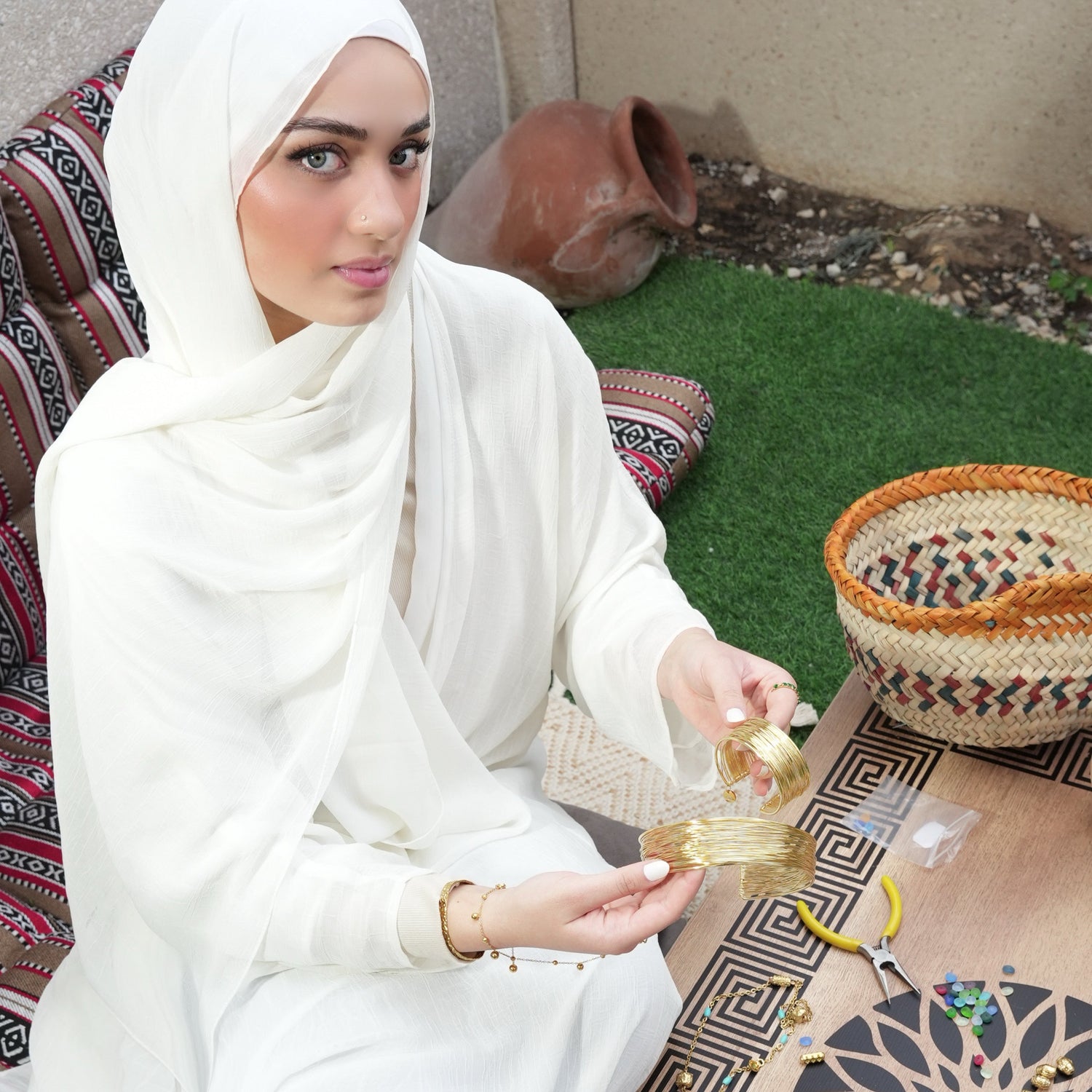 Woman in white traditional attire sitting on a patterned rug with decorative items around her.